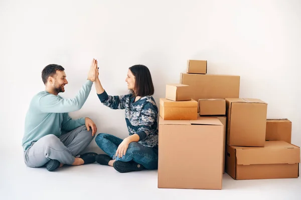 Laughing young couple giving high five sitting on floor in their new home with moving boxes on white background. Success, real estate, moving day concept