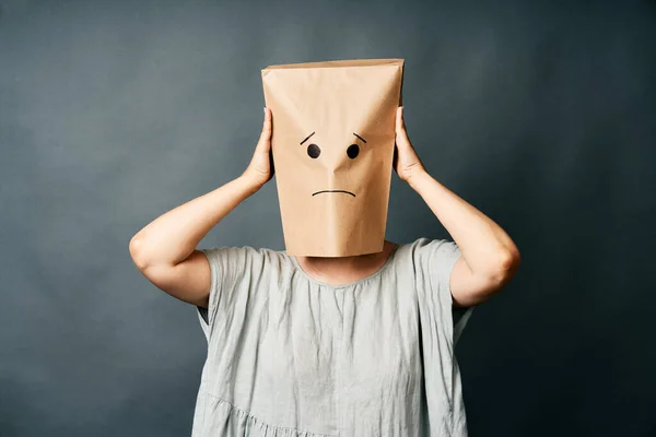 Upset woman with a paper bag on head touching temples, suffering from strong tension headache over gray background. Emotion concept
