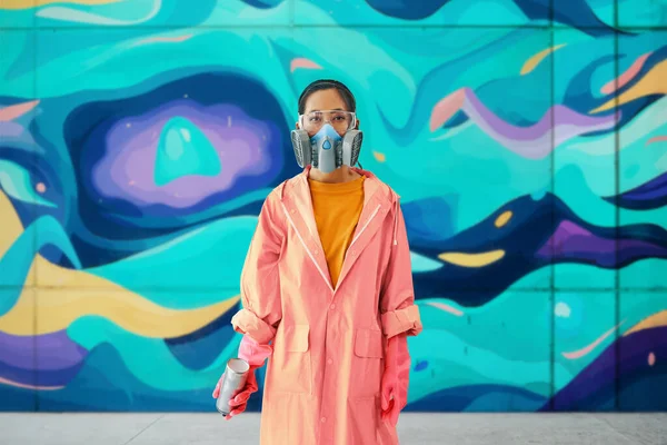 Female graffiti artist in respirator mask standing near the wall with her paintings looking to camera. Street art concept