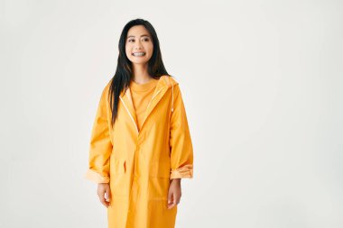 Parlak sarı yağmurluk giyen Asyalı genç gülümseyen kadın beyaz stüdyo arkaplanındaki yeri kopyalamak için kenara bakıyor.
