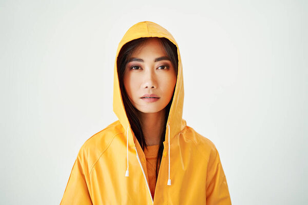 Close up portrait of serious asian girl dressed in bright yellow raincoat posing with hood on her head on white studio background