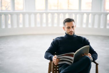 Genç yakışıklı adam dergi okuyor modern beyaz odadaki sandalyede rahatça oturuyordu. Rahatlatıcı kavram