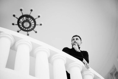 Düşünceli bir adam, klasik beyaz sütunlar ve klasik bir avizeyle balkonda çenesine dokunuyor. Siyah beyaz fotoğraf