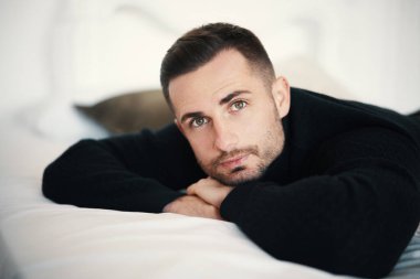 Siyah kazaklı yakışıklı bir adam yatakta dinleniyor, kameraya bakıyor..