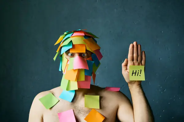 Funny man with cross-eyed covered with colorful sticky notes all over his face and head waving hello over gray background