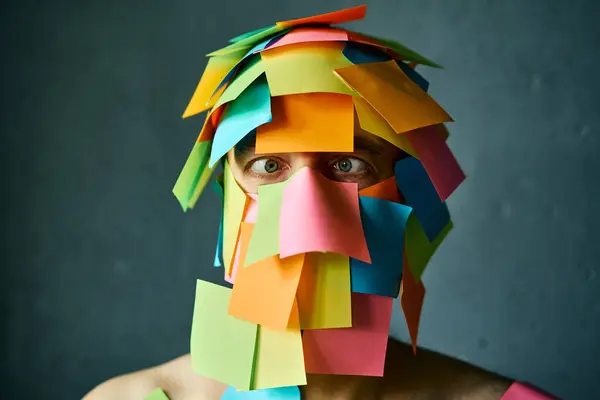 Close up portrait of man with cross-eyed covered with colorful sticky notes all over his face and head over gray background
