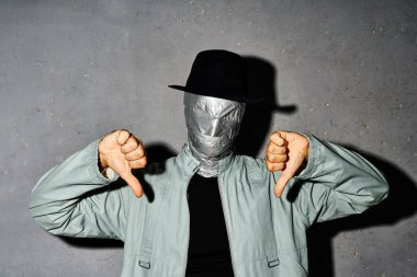 Mystery man with duct tape face showing thumb down sign gesture over gray background clipart