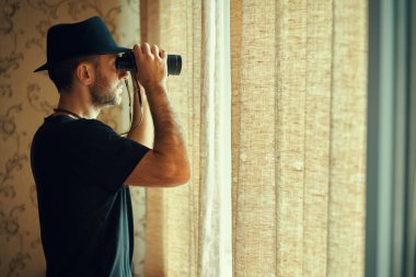 Fötr şapkalı bir adam dürbünle pencereden gözetliyor. Casusluk ve arama kavramı