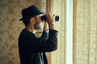 Fötr şapkalı bir adam dürbünle pencereden gözetliyor. Casusluk ve arama kavramı