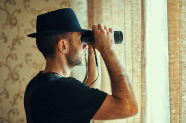 Fötr şapkalı bir adam dürbünle pencereden gözetliyor. Casusluk ve arama kavramı