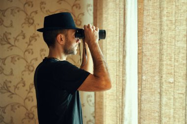 Fötr şapkalı bir adam dürbünle pencereden gözetliyor. Casusluk ve arama kavramı