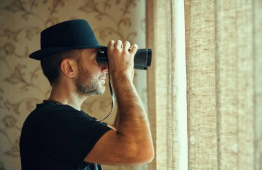 Fötr şapkalı bir adam dürbünle pencereden gözetliyor. Casusluk ve arama kavramı