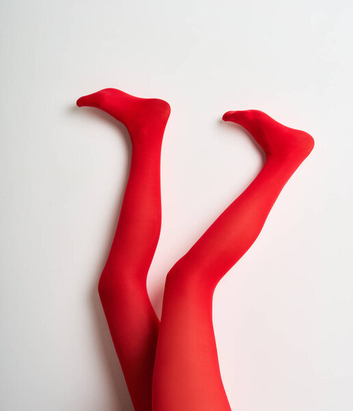 Long slender legs of young woman in red tights raised up posing over white wall background.