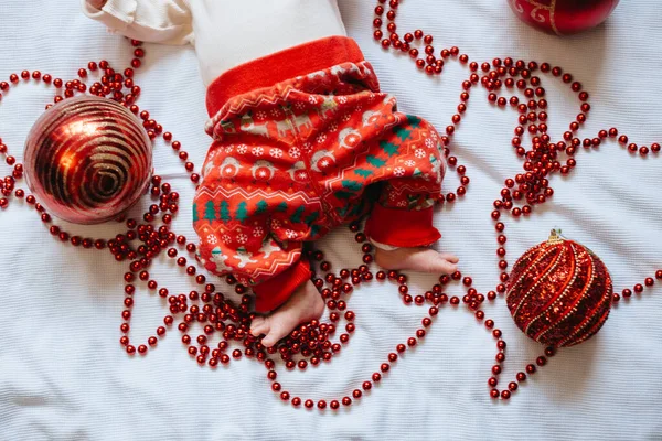 Baby feet in a Christmas garland with Christmas balls. Baby in Christmas pants.