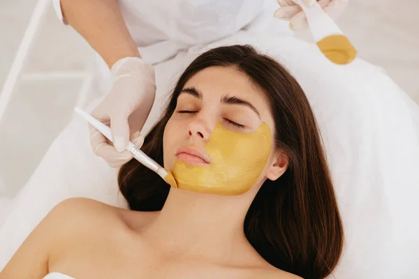 stock image The cosmetologist applies a facial gold mask to the woman's face. Cosmetology and facial skin care in beauty salon. Gold mask. Cosmetic procedure. 