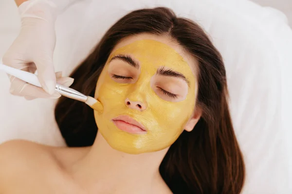 The cosmetologist applies a facial gold mask to the woman\'s face. Cosmetology and facial skin care in beauty salon. Gold mask. Cosmetic procedure.