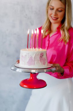 Pembe bluzlu ve beyaz etekli sarışın bir kadın gri arka planda pembe mumlu bir doğum günü pastası tutuyor..