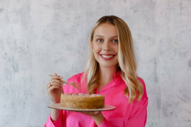 Pembe bluzlu gülümseyen sarışın kadın elinde gri arka planda bir çatal ve cheesecake tutuyor. Mutlu kadın pasta yiyor..