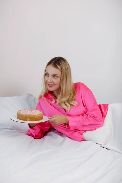 분홍색 블라우스와 치마를 아름다운 금발의 여자가 침대에 하트가 케익을 먹는다 — 스톡 사진