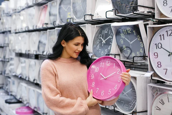 一个穿着粉色毛衣的黑发女人在钟表店里拿着一只粉色的壁表 那个女人身后的墙上有很多钟 — 图库照片