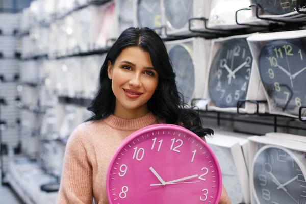 A brunette woman in a pink sweater holds a pink wall watch in a watch store. Lots of wall clocks behind the woman.