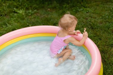 Pembe mayo giyen mutlu bebek yüzüyor ve arka bahçede şişme havuzda oyuncaklarla oynuyor. Yaşam biçimi. 