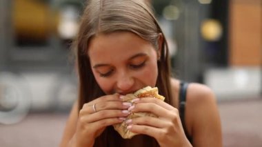 Dışarıda hamburger yiyen, uzun saçlı, bronzlaşmış genç bir kadın. Yaşam tarzı kavramı. 