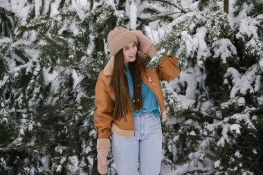 Kahverengi ceketli ve bej şapkalı bir kız karlı bir ormanda çam ağacının yanında..