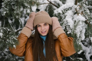 Kahverengi ceketli ve bej şapkalı bir kız karlı bir ormanda çam ağacının yanında..