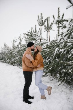 Kahverengi ceketli genç kadın ve adam kışın çamların yanında sarılıyorlar..