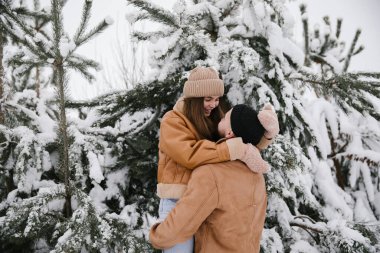 Kahverengi ceketli mutlu çift kışın karlı ormanda eğleniyor. Bir adam bir kadını kollarında tutuyor ve çam ağaçlarının yakınındaki karlı bir ormanda etrafını çeviriyor..