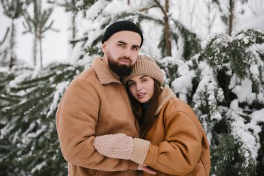 Kahverengi ceketli genç kadın ve adam kışın çamların yanında sarılıyorlar..