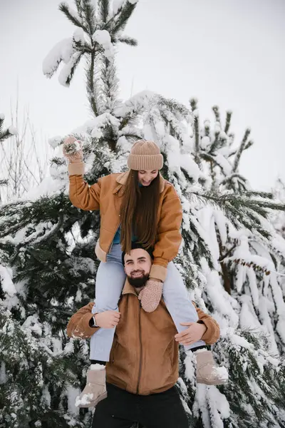 stock image A woman sitting on the shoulders of a man in winter in the snow among snow-covered pines. A couple have fun in a snowy forest in winter.