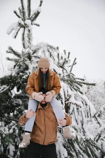 stock image A woman sitting on the shoulders of a man in winter in the snow among snow-covered pines. A couple have fun in a snowy forest in winter.