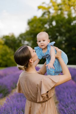 Kahverengi elbiseli anne, küçük kızını lavanta tarlasında mavi elbisesiyle kaldırıyor. Kadın, lavanta tarlasında bir bebeği kaldırıyor..