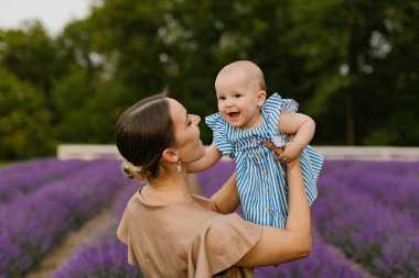 Kahverengi elbiseli anne küçük bebeğini kaldırıyor ve lavanta tarlasında mavi bir elbiseyle çığlık atıyor. Kadın lavanta tarlasında bir bebeği kaldırıyor..