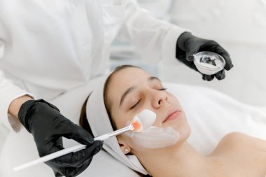 Kozmetik uzmanı sivilceli kadının yüzüne beyaz bir maske takıyor. Gençlik teni. Sivilce derisi. Kozmetikçiyle randevusu olan genç bir kız..