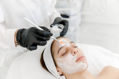 Kozmetik uzmanı sivilceli kadının yüzüne beyaz bir maske takıyor. Gençlik teni. Sivilce derisi. Kozmetikçiyle randevusu olan genç bir kız..