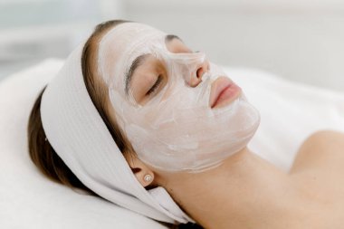 Yüzünde beyaz maske olan genç bir kız kozmetolojist kanepesinde yatıyor. Güzellik salonunda kozmetoloji ve yüz bakımı. Beyaz maske.