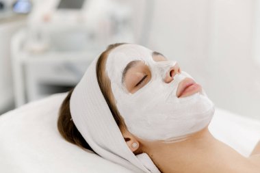 Yüzünde beyaz maske olan genç bir kız kozmetolojist kanepesinde yatıyor. Güzellik salonunda kozmetoloji ve yüz bakımı. Beyaz maske.