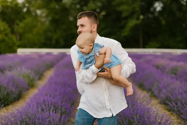 stock image A bearded man in a white shirt is lifting baby in air on a lavender field. Daddy is lifting up daughter and have fun.