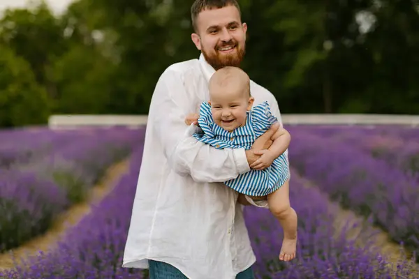 stock image A bearded man in a white shirt is lifting baby in air on a lavender field. Daddy is lifting up daughter and have fun.