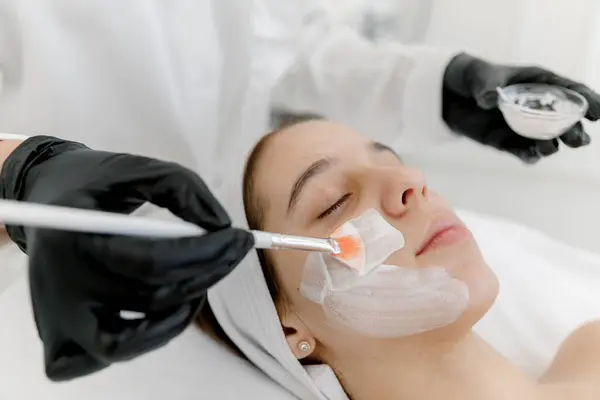 Kozmetik uzmanı sivilceli kadının yüzüne beyaz bir maske takıyor. Gençlik teni. Sivilce derisi. Kozmetikçiyle randevusu olan genç bir kız..