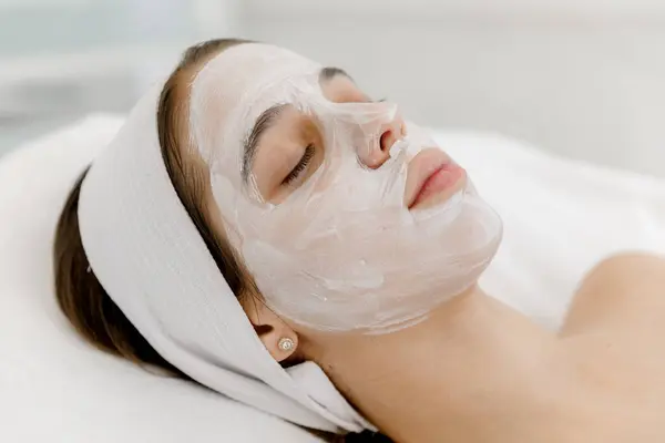 Yüzünde beyaz maske olan genç bir kız kozmetolojist kanepesinde yatıyor. Güzellik salonunda kozmetoloji ve yüz bakımı. Beyaz maske.