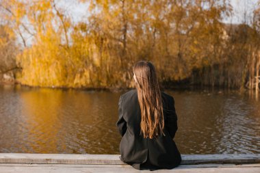 Siyah ceketli, uzun saçlı bir kadın sonbaharda göl kenarındaki ahşap bir duvarda oturuyor. Arka plan.