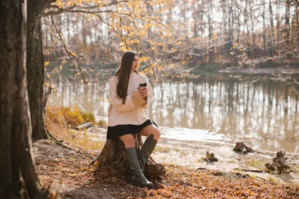 stock image A young brunette in a beige sweater is sitting on a stump and holding a glass of coffee in the forest near the river.