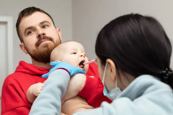 stock image A pediatrician checking the throat of a little baby. Dad is holding a small child in his arms while being examined by a pediatrician.