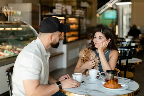 Kahverengi maltipula sahip uzun saçlı bir kadın ve şapkalı sakallı bir adam konuşuyor, kruvasan yiyor ve kafede çay içiyor. Küçük köpeğe aşık bir çift kafede kahvaltı yapıyor..