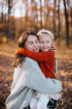 Anne bir sonbahar ormanında kızına saçlarıyla sarılıyor. Gri kazak ve etek giyen kadın sonbahar ormanında kırmızı kazak giyen küçük kızına sarılıyor..