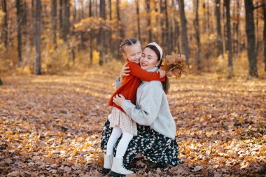 Anne bir sonbahar ormanında kızına saçlarıyla sarılıyor. Gri kazak ve etek giyen kadın sonbahar ormanında kırmızı kazak giyen küçük kızına sarılıyor..